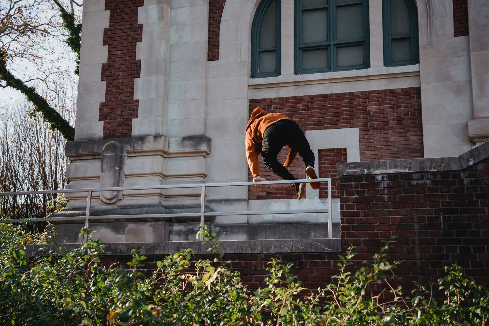 Parkour athlete in an orange hoodie vaulting a metal railing outside a brick building