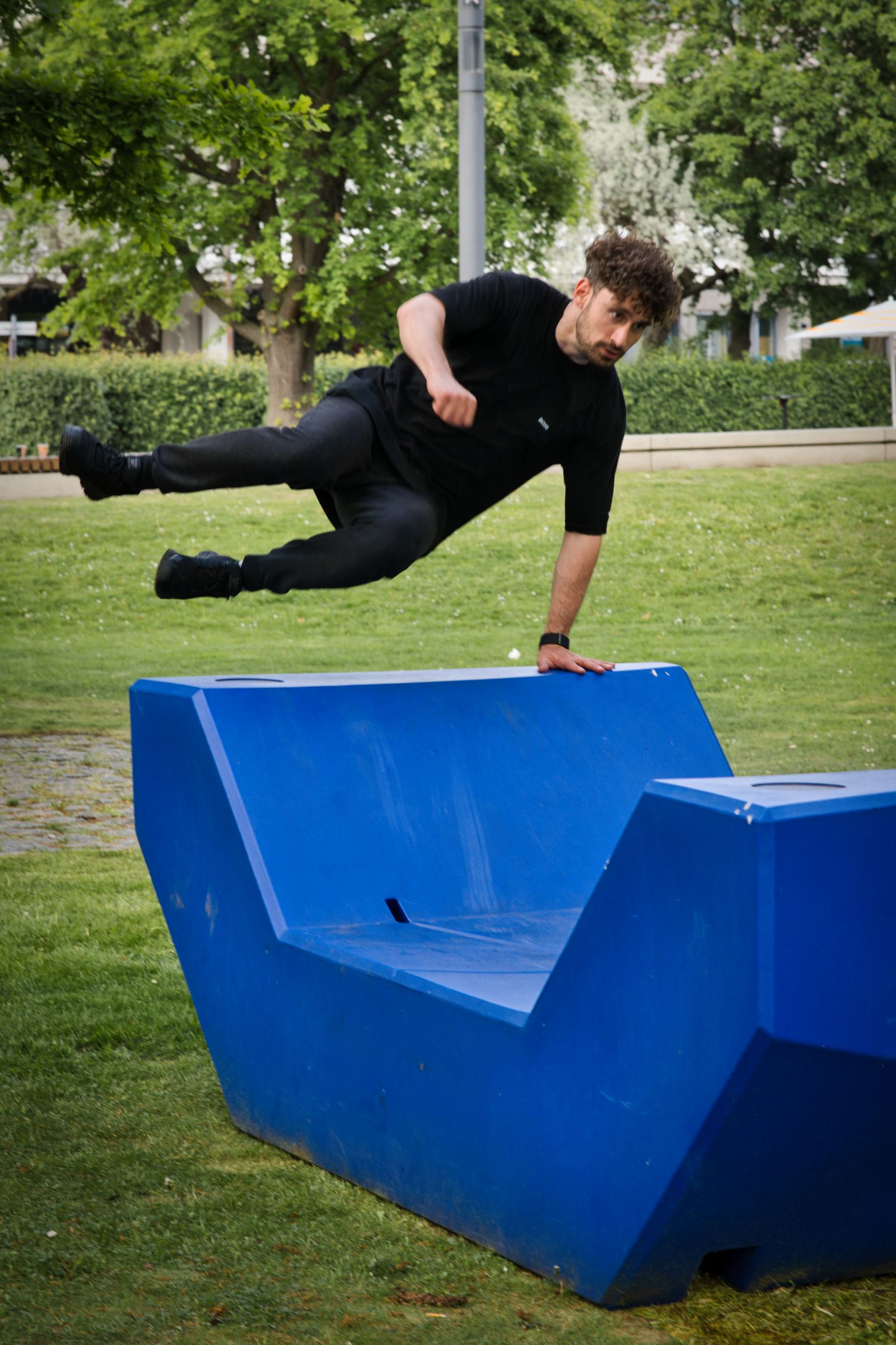 Traceur dressed in black doing a one-handed vault over a blue concrete bench in a park