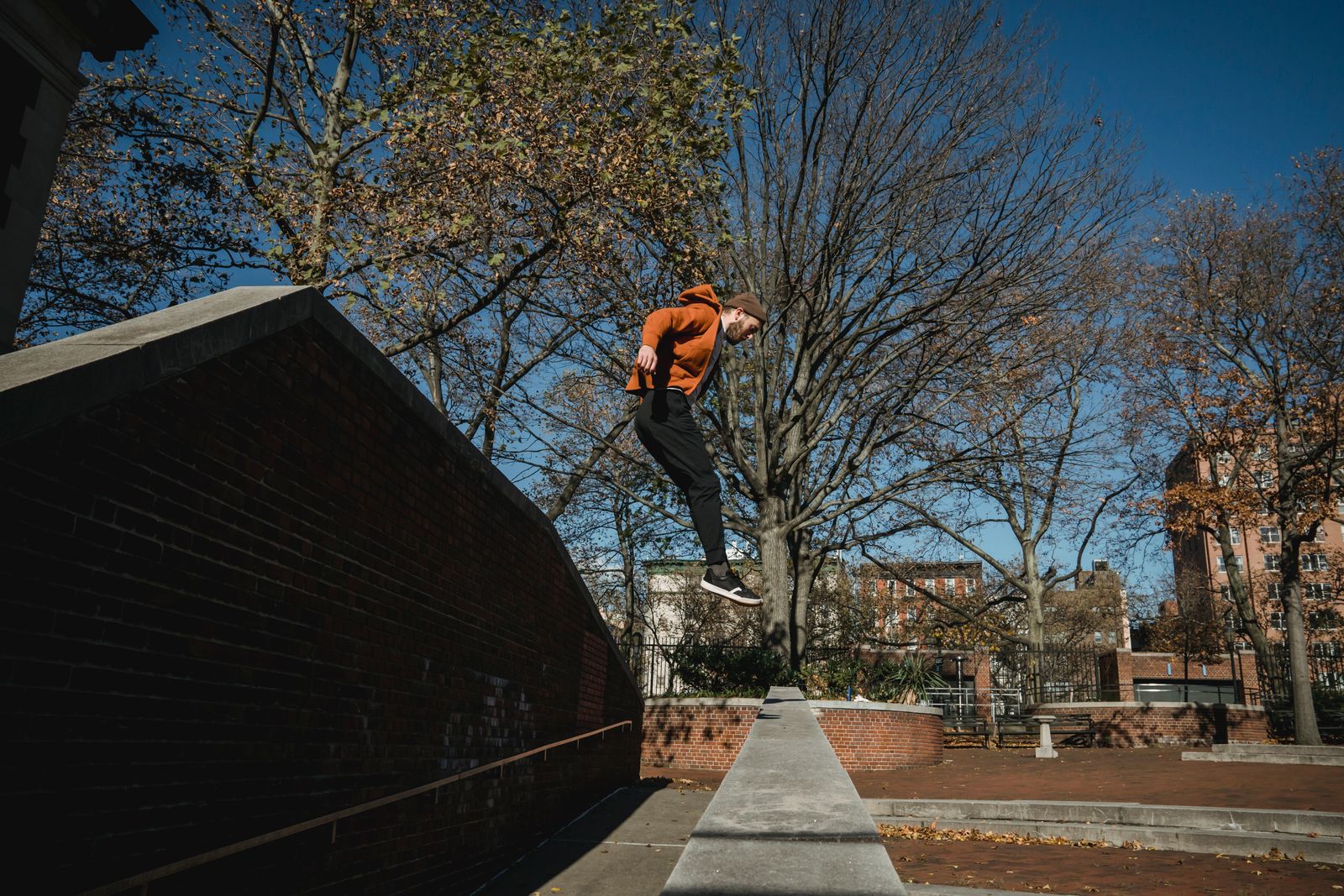 Parkour athlete in an orange hoodie landing a precision jump on a brick wall ledge in an urban park