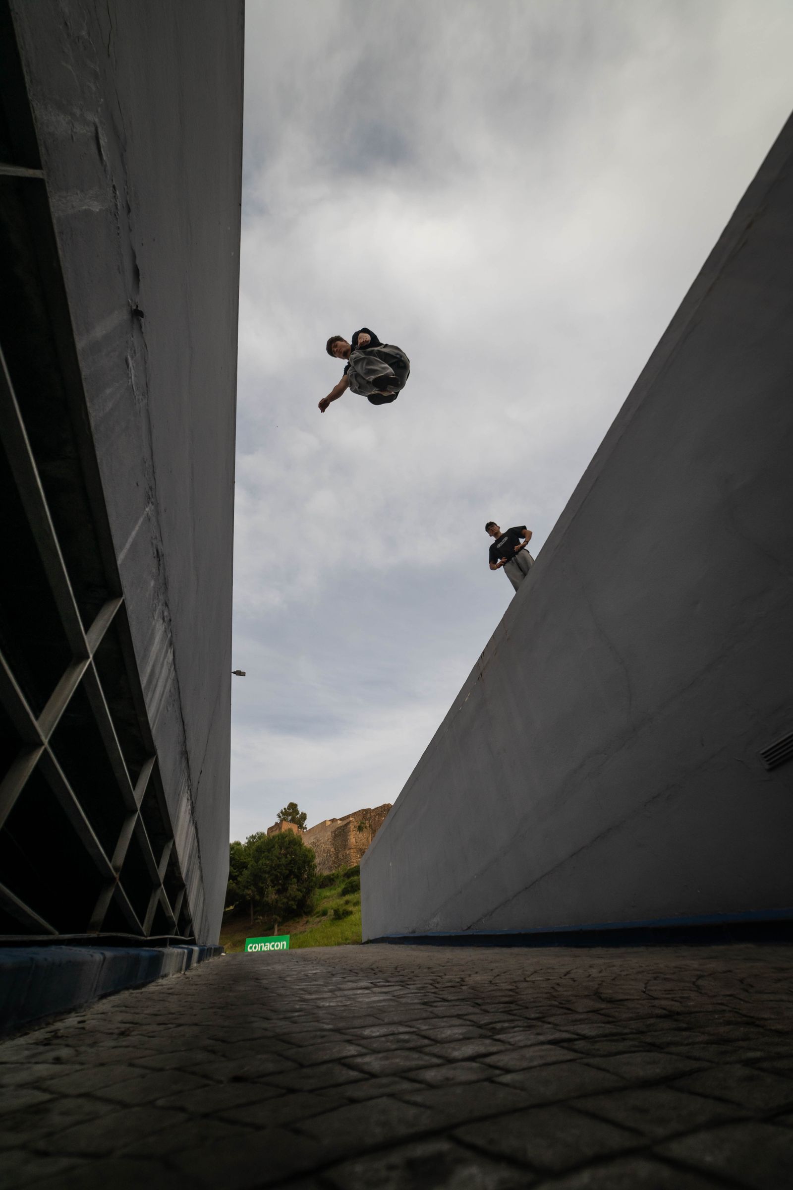 Freerunner performing a high flip between two concrete walls while another athlete watches from a ledge